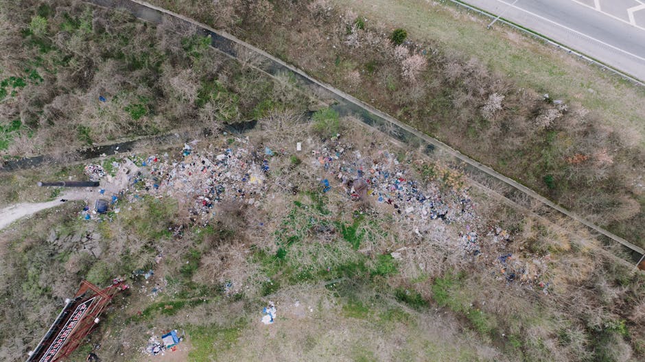 An aerial view showing an extensive area of unmanaged rubbish scattered across a patch of land adjacent to a pathway and surrounded by trees with sparse foliage. The debris consists of various materia