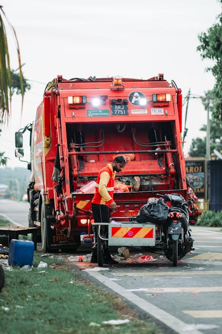 A large, green rubbish bin filled with a mixture of cardboard boxes, some flattened and others still in three-dimensional shape, along with a few white packaging boxes, all showing signs of wear and t